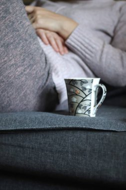 Sıcak bir kanepede oturan bir kadının dikey görüntüsü, yanında duran sıcak bir kahve fincanı. Bulanık. Kişisel bakım, rahatlık ve rahat ev titreşimleri..