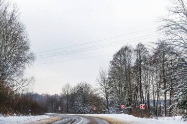 Karla kaplı kış yolu, trafik işaretleriyle kaygan koşullara karşı uyarılıyor. Soğuk ve buzlu atmosfer, kış havasında araba kullanmanın zorluklarını vurguluyor..