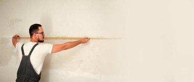 Çalışkan bir inşaat işçisi, bir proje için doğru ölçülerde, temiz beyaz bir duvara karşı mezura kullanır. Bu profesyonel resim geniş kopya alanı olan bir pankart için mükemmeldir.