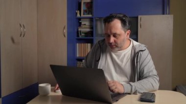 Handsome man working on a laptop computer while sitting at a table at home. Freelancer working from home. Work on the Internet, use of social networks, entertainment in the apartment.