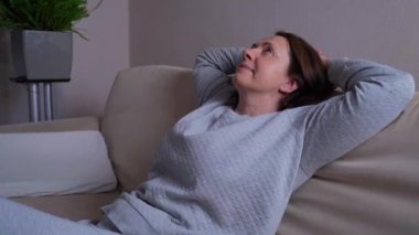 Modern bir oturma odasında, huzurlu Avrupalı bir kadın dinleniyor, kanepeye yaslanıyor, gözlerini kapıyor, ellerini başının arkasına koyuyor, sakin hissediyor, hafta sonunun tadını çıkarıyor.