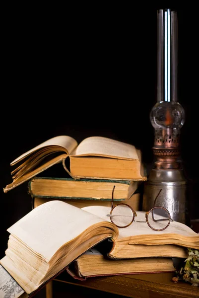 Old antique books with burning paraffin lamp near on the wooden table. Retro style. Vintage still life with old spectacles on book near desk lamp.