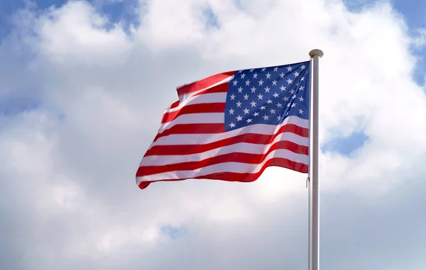Large American flag waving on flag pole with cloud blue sky.