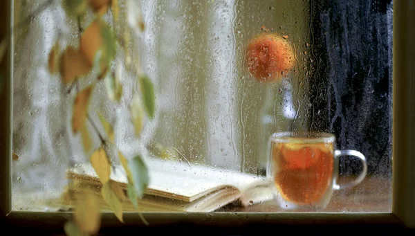 Outside the window, drops of rain flow down the glass. A cup of hot tea, a bouquet of chrysanthemums, an open book
