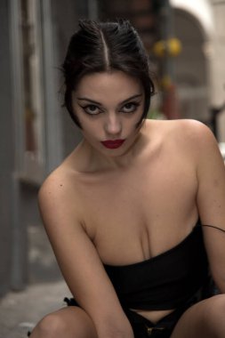 Yoğun makyajlı genç bir kadın şehir ortamında çömelmiş, siyah bir bluz giyiyor.