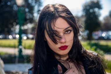 Uzun siyah saçlı, kırmızı rujlu güzel bir kadın güneşli bir günde parkta poz veriyor.