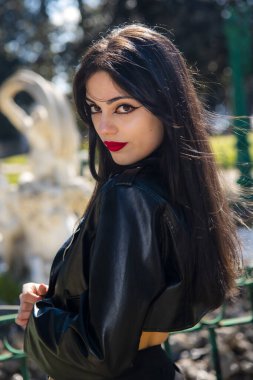 Siyah ceketli, uzun siyah saçlı güzel bir kadın şehir parkında poz veriyor.