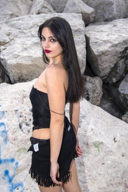 Çekici, uzun siyah saçlı, siyah bluzlu ve etekli bir kadın şehir ortamında poz veriyor.