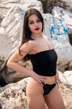 Esmer, uzun saçlı ve kırmızı rujlu bir kadın, bir kaya oluşumunun önünde iç çamaşırıyla poz veriyor.