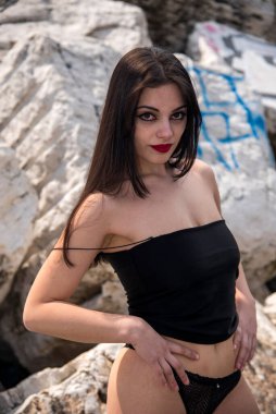 Güneşli bir günde kayalarda poz veren uzun siyah saçlı ve kırmızı rujlu güzel bir genç kadın.