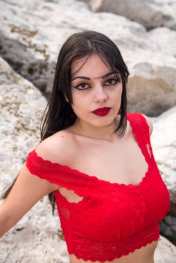 Uzun siyah saçlı ve kırmızı rujlu çekici genç bir kadın bazı kayalarda poz veriyor.