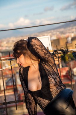 Şehvetli bir kadın gün batımında çatıda poz veriyor. Siyah dantelli bir elbise giyiyor. Arka planda gökyüzü var.