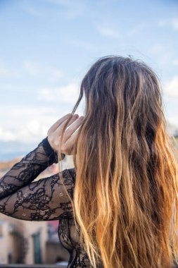 Uzun saçlı genç bir kadın saçlarına dokunuyor ve mavi gökyüzünün altındaki manzaraya bakıyor.