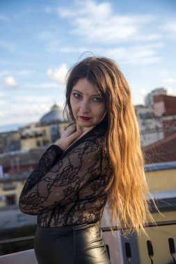 Güneşli bir günde şehrin çatısında şık poz veren kendine güvenen bir manken. Uzun kızıl saçları zarif bir şekilde süzülüyor ve modaya uygun siyah deri dantelli bir elbiseyle şehvet ve cazibe yayıyor.