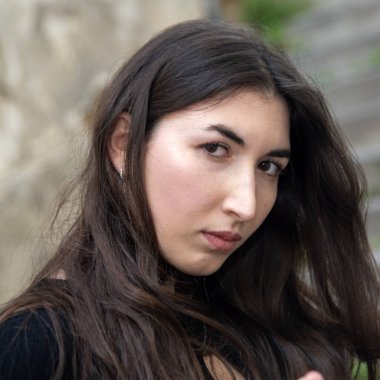 Uzun kahverengi saçlı, ciddi bir genç kadın dışarıda poz veriyor, büyüleyici bir portre oluşturuyor.