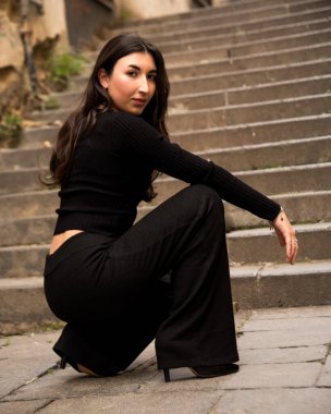 Siyah kıyafetli, şık bir genç kadın şehir ortamında taş basamaklara çömelmiş şık ve anımsatıcı bir portre çiziyor.