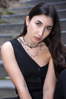 Uzun siyah saçlı, şık aksesuarları olan şık bir genç kadın şehir ortamında taş merdivenlerde oturuyor.