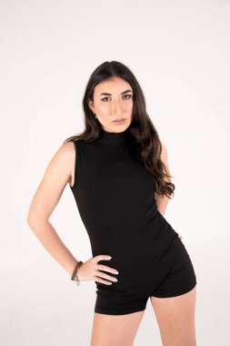 Tarz sahibi genç bir kadının, beyaz bir arkaplanda, şık, siyah bir elbise giymiş, kendine güvenen bir şekilde poz verdiği bir fotoğraf.