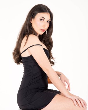 Genç esmer manken, dar siyah bir elbise giymiş, beyaz bir arka planda şık bir stüdyo çekimi için poz veriyor.