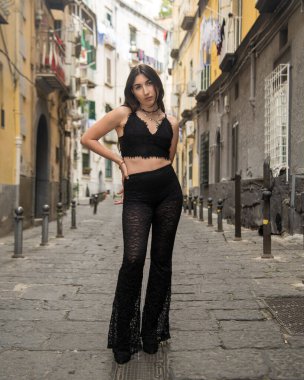 Napoli sokaklarında poz veren uzun kahverengi saçlı genç bir kadın siyah bluz ve dantel pantolon giyiyor.