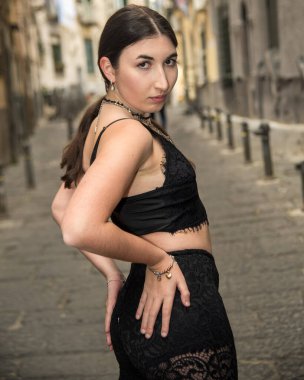 Şık genç bir kadın dar bir sokak arasında kendine güvenen bir duruş sergiliyor modern moda ve şehrin arka planına karşı tavır sergiliyor.