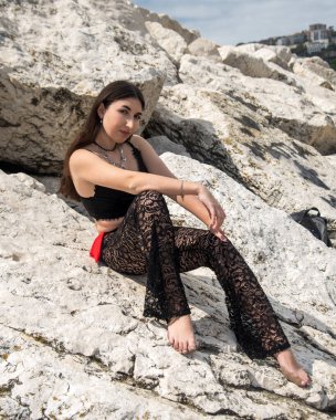 Deniz kenarındaki büyük kayalarda oturan kadın güneşli bir günün tadını çıkarıyor. Şık dantel pantolon ve siyah bir bluz giyiyor, rahat bir tavır sergiliyor.