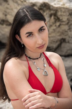 Kalp şeklinde bir kolye takan ve canlı kırmızı bir bikiniyle kayalık bir sahilde poz veren modaya uygun bir manken.