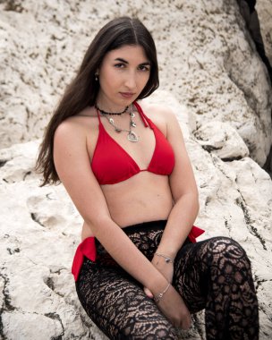 Uzun kahverengi saçlı, kırmızı bikinili ve siyah balıkçı pantolonlu, büyük beyaz bir kayanın üzerinde oturan şık bir genç kadın.