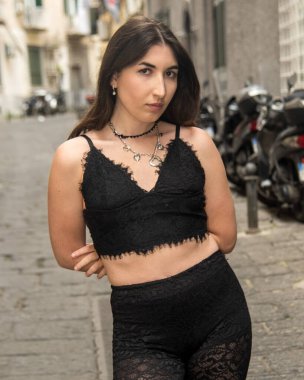 Dar bir sokakta şık genç bir kadın, modaya uygun siyah dantelli bir kıyafet giyiyor, kendine güveni ve tarzı var.