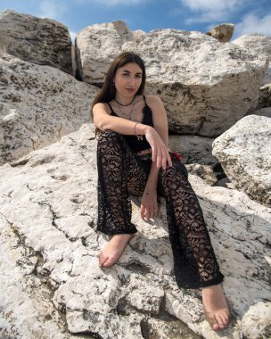 Beyaz kayaların üzerinde çıplak ayakla poz veren manken siyah dantelli pantolon ve mavi gökyüzü güneşli bir günde siyah bluz giyiyor.