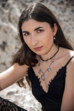 Uzun kahverengi saçlı, siyah dantelli ve gümüş kolyeli, beyaz taşların üzerinde oturan zarif bir genç kadın.