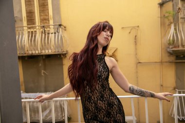 Siyah şeffaf elbiseli, balkonda şehvetli pozlar veren kızıl saçlı bir manken.