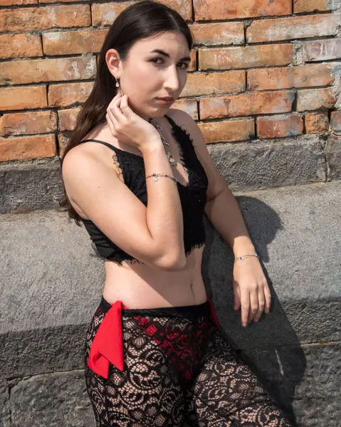 Dantelli elbiseli, zarif bir kadın. Tuğla ve taş bir duvara karşı güvenle poz veriyor. Modern stil ve zarafet sergiliyor.