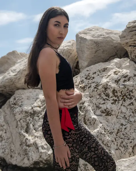 Siyah dantelli bluz ve kırmızı kuşaklı taytlı beyaz kayaların üzerinde poz veren manken.