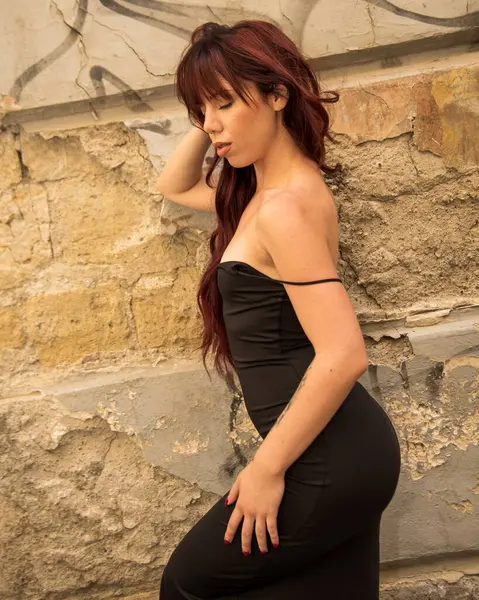 Kızıl saçlı manken dar siyah bir elbise giyip eli saçında ve gözleri kapalı, yıpranmış eski bir duvarın yanında poz veriyor.