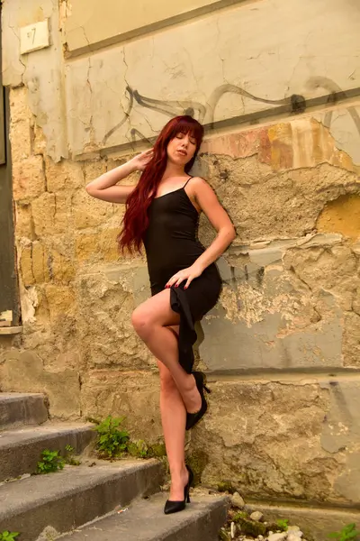 Uzun kızıl saçlı, dar siyah elbiseli ve şehir ortamında açık merdivenlerde siyah topuklu giyen manken.