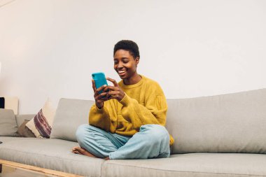 African woman using cellphone at home sitting on the sofa