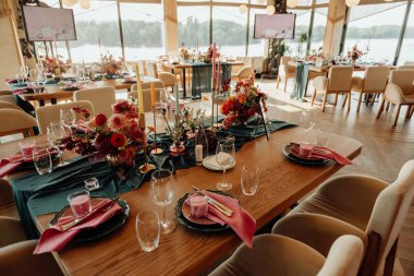 Decoration and decoration of wooden tables in a restaurant for a holiday. Professional decoration, serving and floristry with fresh flowers. Decoration with flowers and candles in turquoise and red colors, decoration elements in glass and fabric in g