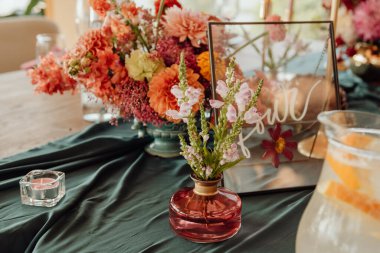Detail of a table setting in a restaurant. Fresh flowers in a small vase on a green tablecloth. Next to the flowers is a candle in a glass candlestick. Against the background of a large bouquet of flowers of red and orange flowers