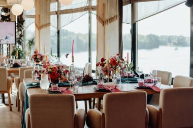 A table in a restaurant on the river bank with panoramic windows, professionally decorated by decorators and florists. On the table are details of glass and crystal, fresh flowers of red and orange, candles in candlesticks