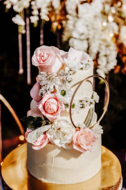 El yapımı bir düğün pastası süslenmiş bir düğün fotoğrafının arka planında altın bir tepside duruyor. Gece çekimi. Pasta pembe güller, beyaz papatyalar ve diğer çiçeklerle süslenmiş..