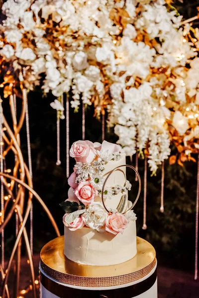 A handmade wedding cake stands on a golden tray against the backdrop of a decorated wedding photo zone. Night shooting. The cake is decorated with pink roses, white daisies and other flowers.