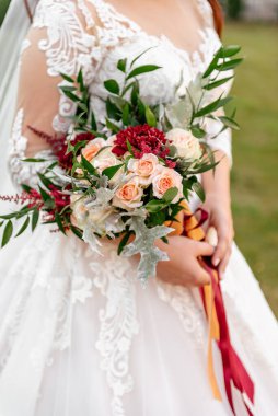 Bir buket kırmızı kasımpatı ve yeşil yapraklar ve kırmızı kurdelelerle süslenmiş pembe güller, dantelli, bulanık bir arka planda, beyaz elbiseli bir gelinin ellerinde.