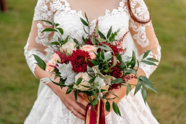 Bir buket kırmızı kasımpatı ve yeşil yapraklar ve kırmızı kurdelelerle süslenmiş pembe güller, dantelli, bulanık bir arka planda, beyaz elbiseli bir gelinin ellerinde.