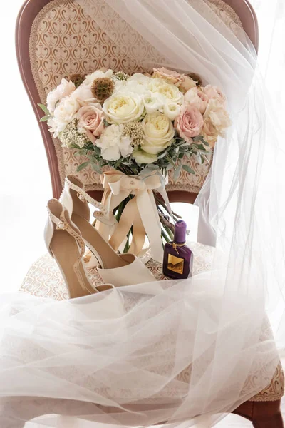 stock image On a classic wooden expensive chair with a white veil laid out on it, there is a bouquet of fresh flowers, the bride's shoes and a bottle of perfume close-up