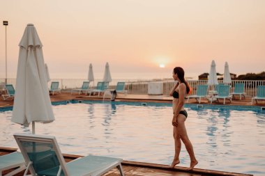 Mayo giymiş ıslak bir kız açık havuzun kenarında duruyor. Günbatımında insanlar olmadan