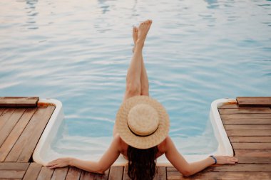 Hasır şapkalı bir kız açık havuza inerken ellerini havuzun kenarına yaslıyor. O, bacaklarını onun önünde düzleştirdi. Kızın bacaklarına odaklan.