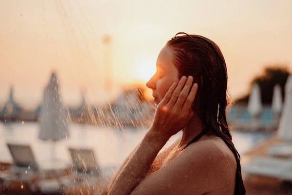 The girl washes her face with water in the shower, water pours on her face, she covers her face with her hands. Sunset and outdoor pool in the background