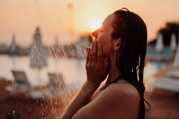 The girl washes her face with water in the shower, water pours on her face, she covers her face with her hands. Sunset and outdoor pool in the background