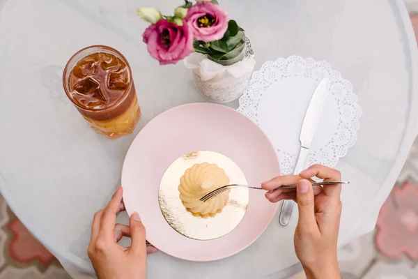 The girl takes a dessert with a fork that lies on a golden plate. Hand holds a glass with a drink in which ice floats. The table is decorated with a white tablecloth and served with cutlery
